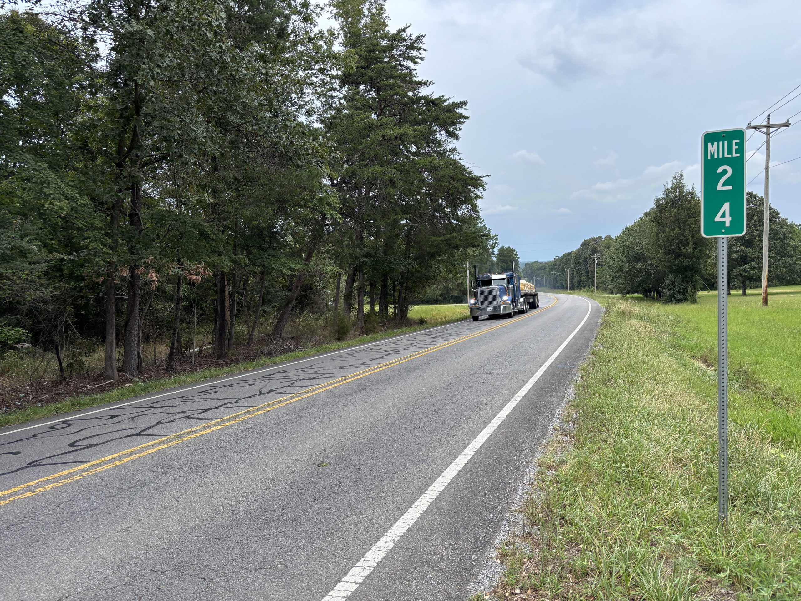 A tractor trailer travels on a two-lane highway, approaching a 24 mile marker on the road's shoulder.