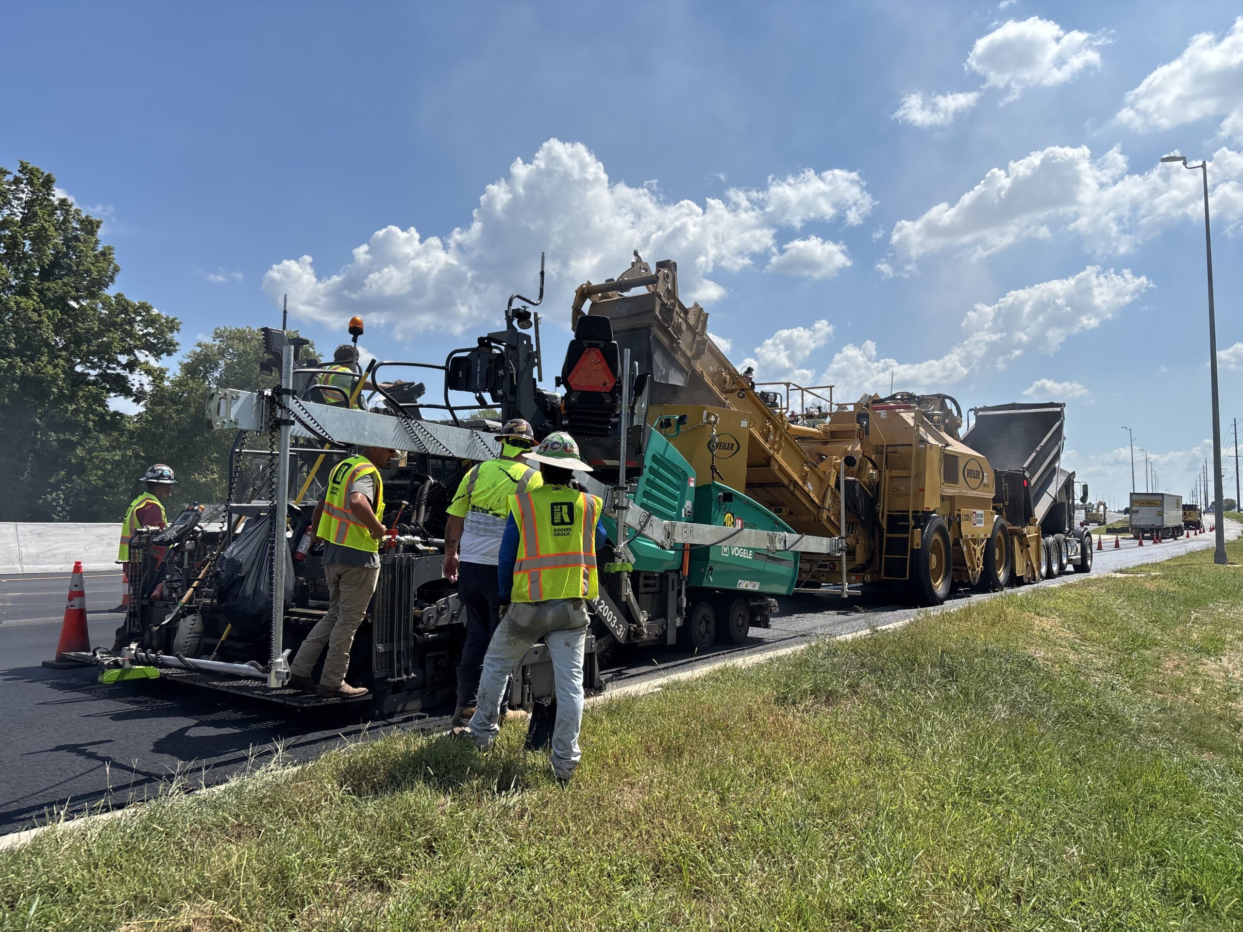 Workers move alongside a paving train consisting of a paver, material transfer vehicle and dump trucks delivering asphalt.