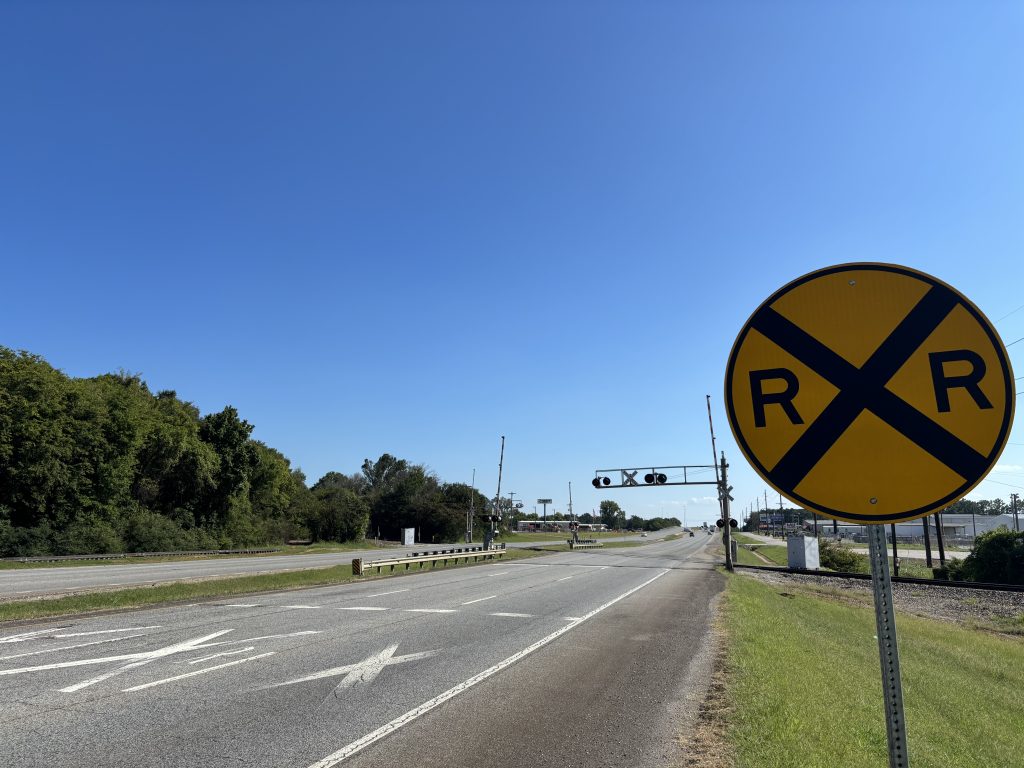 A railroad warning sign ahead of a rail crossing with gates and signals on a divided six-lane highway.