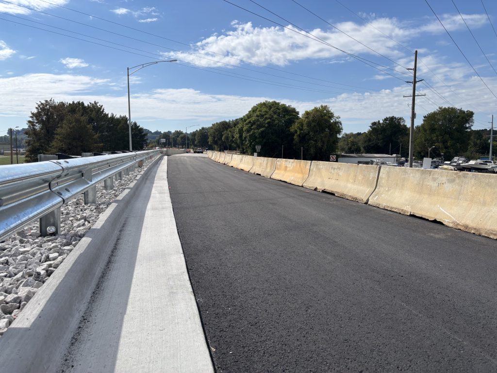 A new concrete bridge at the end of a new asphalt road lined by new curb, gutter and guardrail on one side and concrete barriers on the other.