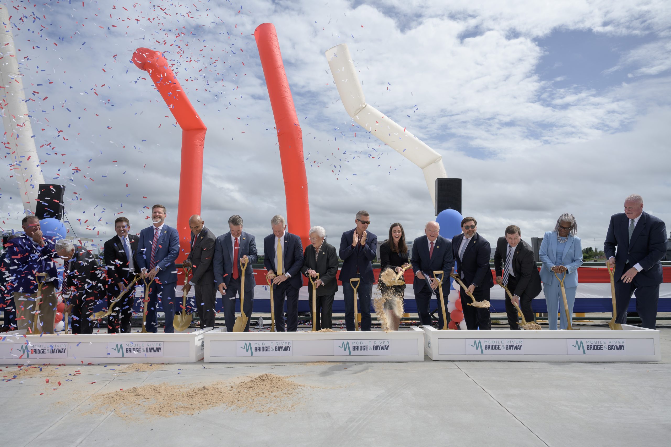 Groundbreaking participants turn soil with gold shovels behind boxes displaying the Mobile River Bridge and Bayway logo as confetti falls.